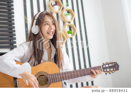 Portrait young smile happy beautiful asian woman relax bedroom. woman with headphones playing acoustic guitar in white bedroom Portrait young smile happy beautiful asian woman relax bedroom. woman with headphones playing acoustic guitar in white bedroom 88958431