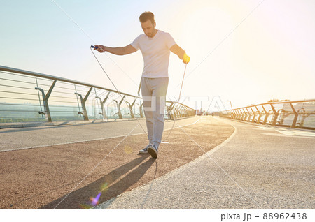Full length portrait of a muscular build European middle aged man, fit athlete doing jumping exercises, cardio training with skipping rope on the urban city bridge early in the morning on a summer day 88962438