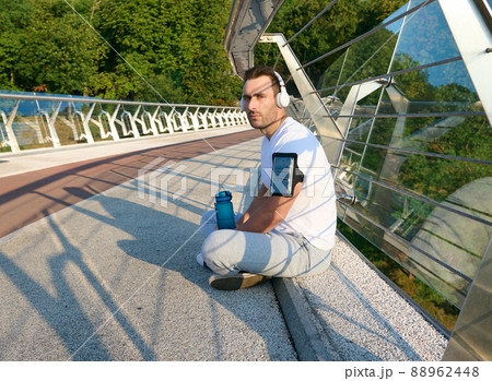 Confident relaxed handsome European man, athlete in sportswear and wireless headphones, listening to the music while relaxing on the urban glass bridge after morning workout on a warm summer sunny day Confident relaxed handsome European man, athlete in sportswear and wireless headphones, listening to the music while relaxing on the urban glass bridge after morning workout on a warm summer sunny day 88962448