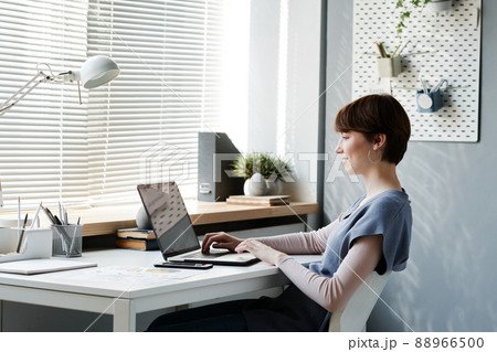 Smiling young female strategic manager sitting at desk and browsing internet on laptop while working on project online 88966500