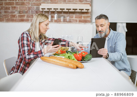 Mature couple sitting at kitchen table with gadgets in hands Mature couple sitting at kitchen table with gadgets in hands 88966809