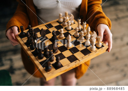 High-angle view cropped shot of unrecognizable young woman holding in hands chess board standing on vintage wooden floor in dark room, selective focus, close-up. High-angle view cropped shot of unrecognizable young woman holding in hands chess board standing on vintage wooden floor in dark room, selective focus, close-up. 88968485