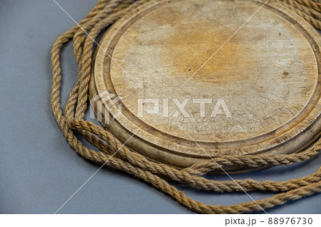 Ship's rope wrapped around a cutting board. Round empty wooden cutting board and ship's rope against a gray background. View from above at an angle. Close-up. Selective focus. Ship's rope wrapped around a cutting board. Round empty wooden cutting board and ship's rope against a gray background. View from above at an angle. Close-up. Selective focus. 88976730