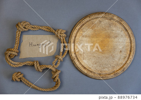 Round empty wooden cutting board against a gray background. Ship's rope with knots. Scrap of paper with handwritten text HUNGRY? Top view 88976734