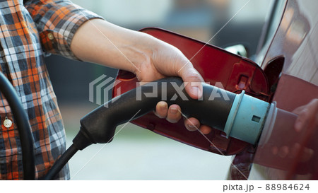 Female model stands by an electric car and holds a charging plug Female model stands by an electric car and holds a charging plug 88984624