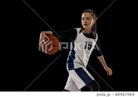 Training. Studio shot of teen girl in white uniform playing basketball, doing dribbling exercise isolated over black background. Training. Studio shot of teen girl in white uniform playing basketball, doing dribbling exercise isolated over black background. 88987069