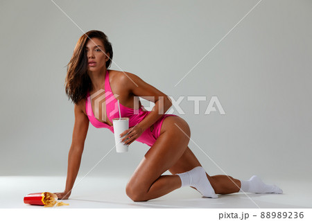 cheat meal concept. athlete woman is posing with fast food on light gray background, studio shot. young sporty woman is holding fast food close up. cheat meal concept. athlete woman is posing with fast food on light gray background, studio shot. young sporty woman is holding fast food close up. 88989236