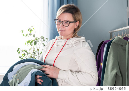 Real middle aged millennial woman seamstress holding suits sewn from multicolored fabric footer. Tailor taking clothes off hangers. Hand making clothes in home interior 88994066