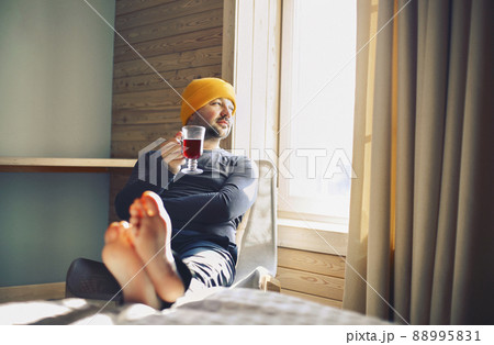 Winter male traveller in yellow hat holds a mug of mulled wine with by the window in wooden cozy house Winter male traveller in yellow hat holds a mug of mulled wine with by the window in wooden cozy house 88995831