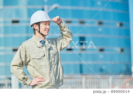 ビルの前にヘルメットをかぶって立つ作業服姿の若い男性 89000014