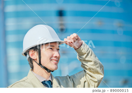 ビルの前にヘルメットをかぶって立つ作業服姿の若い男性 89000015