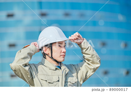 ビルの前でヘルメットをかぶる作業服姿の若い男性 89000017