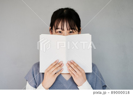 Portrait of Asian student girl with bangs covering face behind book against isolated background 89000018