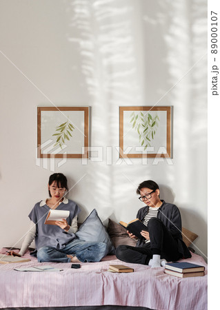 Focused Asian students sitting on bed and reading book and workbook before exam 89000107