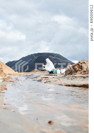 Environmental engineer in protective suit crouching at river and taking sample of water while checking contaminated water Environmental engineer in protective suit crouching at river and taking sample of water while checking contaminated water 89000612