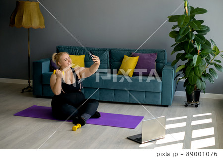 An obese young blonde woman with a dumbbell takes a selfie on her smartphone during a home workout. Sports at home in the living room with a green sofa and yellow-purple pillows. Model plus size 89001076