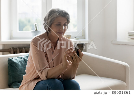 Attractive older female holding smartphone make call seated on sofa 89001861