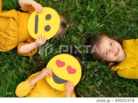 Three funny girlfriends are lying on the green grass with different emoticons in their hands. One girl is laughing, and two are holding smile masks covering their faces. Types of temperament. 89002478