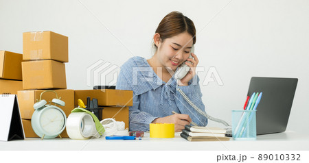 Young Asian woman business owner with many parcel boxes on the table happy online sales job, use the laptop, get an order from customers, take notes, and make arrangements for delivery by post 89010332