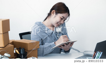 Young Asian woman business owner with many parcel boxes on the table happy online sales job, use the laptop, get an order from customers, take notes, and make arrangements for delivery by post 89010333