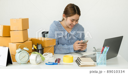 Young Asian woman business owner with many parcel boxes on the table happy online sales job, use the laptop, get an order from customers, take notes, and make arrangements for delivery by post 89010334