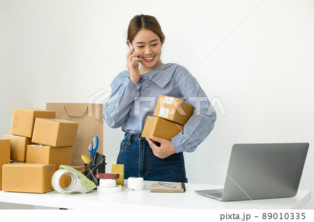 Young Asian woman business owner with many parcel boxes on the table happy online sales job, use the laptop, get an order from customers, take notes, and make arrangements for delivery by post 89010335