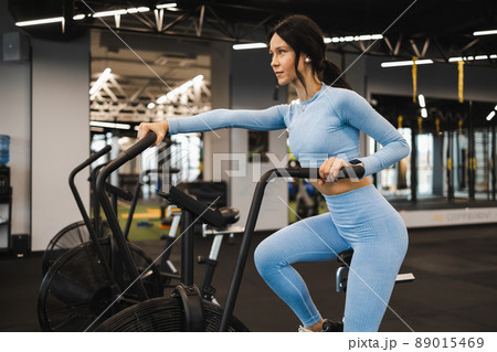 Woman riding on air bike in gym. .Girl using exercise bike for cardio training in the crossfit studio Woman riding on air bike in gym. .Girl using exercise bike for cardio training in the crossfit studio 89015469