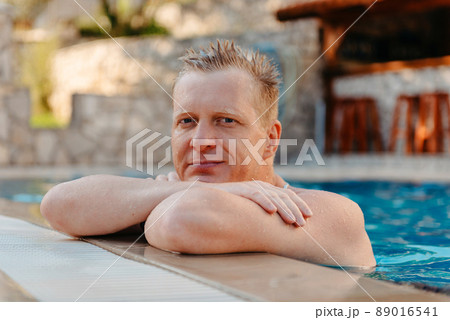 Photo of handsome smiling man in swimming pool in summer scenery. Attractive, cheerful, stunning, perfect, harsh, virile man in yellow shorts, having cocktail with small umbrella in hand and holding 89016541