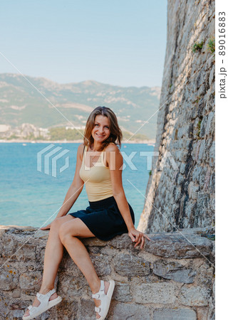 Beautiful girl sitting on a stone wall, in background is the blue sea, Budva, Montenegro. 89016883
