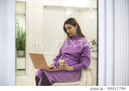 Beautiful brunette pregnant woman with glass of milk and using a laptop online sitting on the chair near window in light room at home 89017796