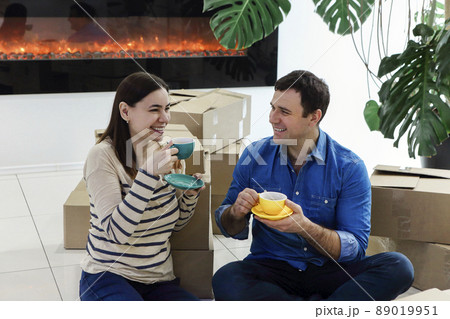 Young mixed ethnicity couple taking a break on moving day into new home sitting on floor in lounge drinking coffee surrounded by removal boxes by the fireplace 89019951