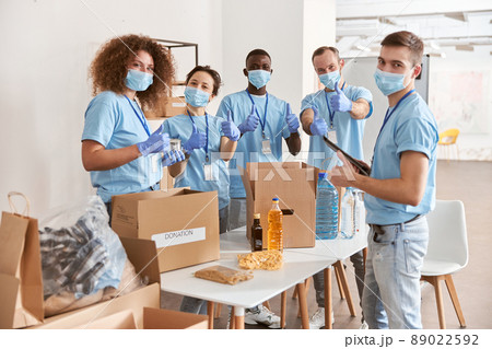 Group of diverse people wearing blue uniform, protective masks and gloves showing thumbs up while sorting donated food items, volunteering in community together 89022592