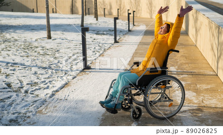 Happy woman in wheelchair happily pulls her hands up while walking in winter park. Happy woman in wheelchair happily pulls her hands up while walking in winter park. 89026825