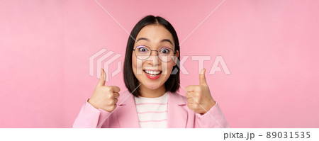 Awesome, congrats. Face of excited asian businesswoman in glasses, smiling pleased, showing thumbs up in approval, standing over pink background 89031535