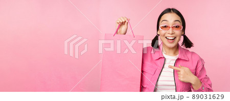 Shopping. Stylish asian girl in sunglasses, showing bag from shop and smiling, recommending sale promo in store, standing over pink background Shopping. Stylish asian girl in sunglasses, showing bag from shop and smiling, recommending sale promo in store, standing over pink background 89031629