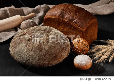 Variety of baked bread and buns with sesame and sunflower seeds, sprinkled with flour. Bunch of wheat, burlap, rolling pin, black background. Close-up 89032219