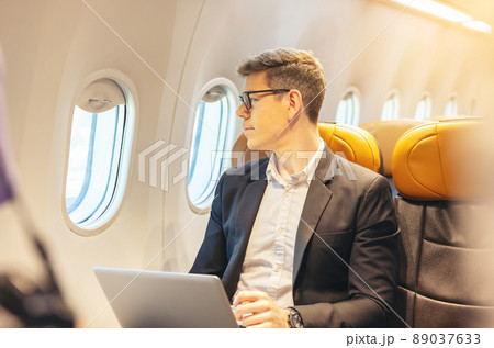 During a flight, a businessman in formal attire and glasses looks out the plane window while working on a laptop computer. Service with Internet access on board and a business travel concept 89037633