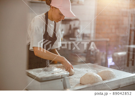 Woman in pink cap kneads raw dough piece for bread in craft bakery shop Woman in pink cap kneads raw dough piece for bread in craft bakery shop 89042852