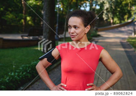 Charming muscular woman with healthy aesthetic body and glowing skin, athlete in red t-shirt and smartphone holder posing with arms on waist looking aside standing on the city park treadmill Charming muscular woman with healthy aesthetic body and glowing skin, athlete in red t-shirt and smartphone holder posing with arms on waist looking aside standing on the city park treadmill 89062824