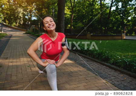 Happy cheerful African American woman doing lunges while exercising outdoor, smiles cutely with beautiful toothy smile looking at camera, enjoying her active and healthy lifestyle 89062828