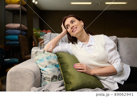 Beautiful woman sitting on a new sofa on sale, examining it before buying. Attractive woman shopping for upholstered furniture at shopping mall. Consumerism, interior design and new home concept 89062941