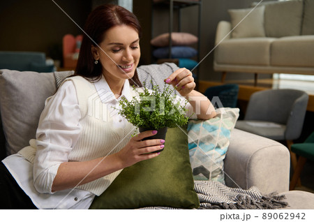 Beautiful middle-aged woman holding ornamental plants sitting on a stylish modern comfortable sofa while shopping in the upholstered furniture store. Interior Design and home improvement 89062942