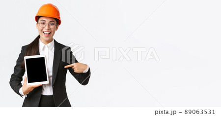 Smiling professional female construction manager, asian engineer in safety helmet and business suit show project, pointing finger at digital tablet screen with pleased expression, white background Smiling professional female construction manager, asian engineer in safety helmet and business suit show project, pointing finger at digital tablet screen with pleased expression, white background 89063531