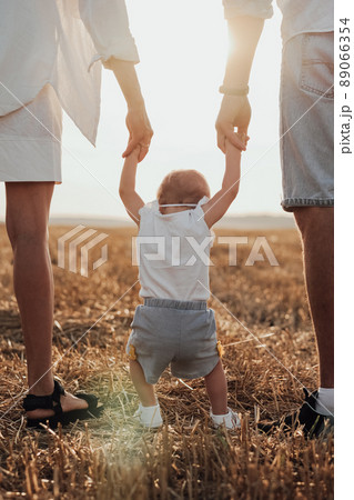 Close Up Back View of Mom and Dad Helping Their Child Making First Steps, Young Family Walking on the Field at Sunset Close Up Back View of Mom and Dad Helping Their Child Making First Steps, Young Family Walking on the Field at Sunset 89066354