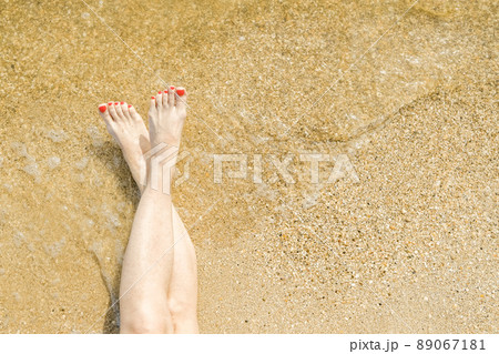 Top view of beautiful female feet with bright red pedicure on the sand of the beach. The sea wave washes women's feet. Relaxation and enjoyment during your seaside holiday. Copy space. 89067181