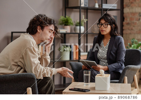 Horizontal medium shot of sad young man treating depression with help of professional psychotherapist 89068786