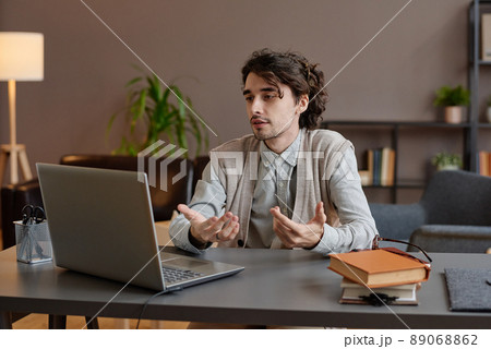 Horizontal medium portrait of young adult psychotherapist wearing casual outfit sitting at desk in his office on video call using laptop Horizontal medium portrait of young adult psychotherapist wearing casual outfit sitting at desk in his office on video call using laptop 89068862
