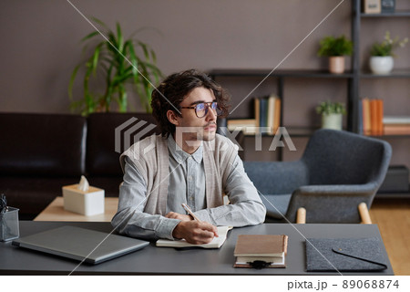 Horizontal medium portrait of handsome young adult man wearing stylish casual outfit with eyeglasses sitting at desk looking away 89068874