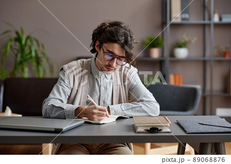 Horizontal medium portrait of young adult man wearing comfortable casual outfit and eyeglasses sitting at desk writing something in notebook 89068876