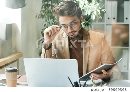 Busy young middle eastern businessman in beige jacket adjusting eyeglasses while working with website statistics on laptop 89069368